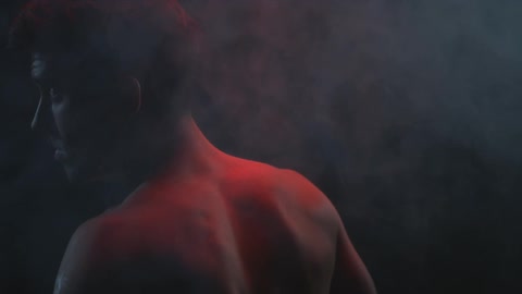 A man stands in the midst of a smoky room lit from above by a red light