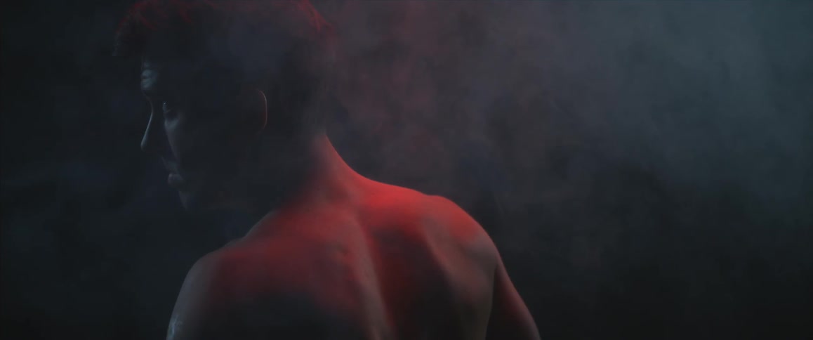 A man stands in the midst of a smoky room lit from above by a red light