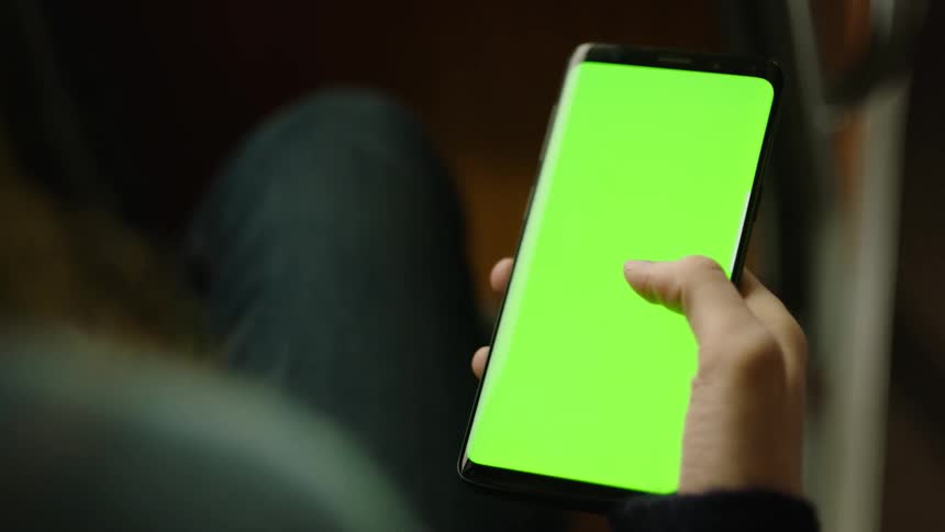 Close-up of a person sitting on a bus holding a smartphone with a green screen and repeatedly swiping up with their thumb. Perfect for VFX, screen replacement, app mockups, UI design, and mobile interaction scenes in everyday commuting contexts.