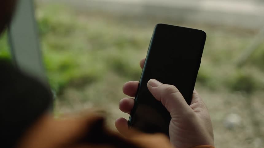 Close-up of a person sitting at a bus stop holding a smartphone with a dark screen and swiping multiple times from left to right with their thumb. Perfect for VFX, screen replacement, UI design, mobile app mockups, and modern commuting scenes.