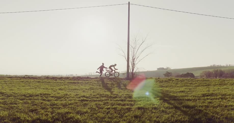 Cyclist rides a bike down a country road while a filmmaker running tries to keep up and falls down