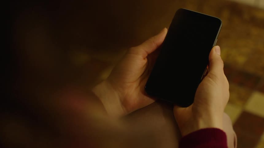 Cinematic close-up of a person holding a smartphone with a black screen indoors under warm light. The phone stays still without interaction, perfect for screen replacement, VFX, compositing, app mockups, and modern digital lifestyle projects.