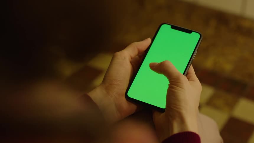 Cinematic close-up of a person sitting indoors, holding a smartphone with a green screen in both hands and typing with both thumbs. Filmed in warm light, perfect for VFX, screen replacement, app mockups, messaging, and digital communication projects.