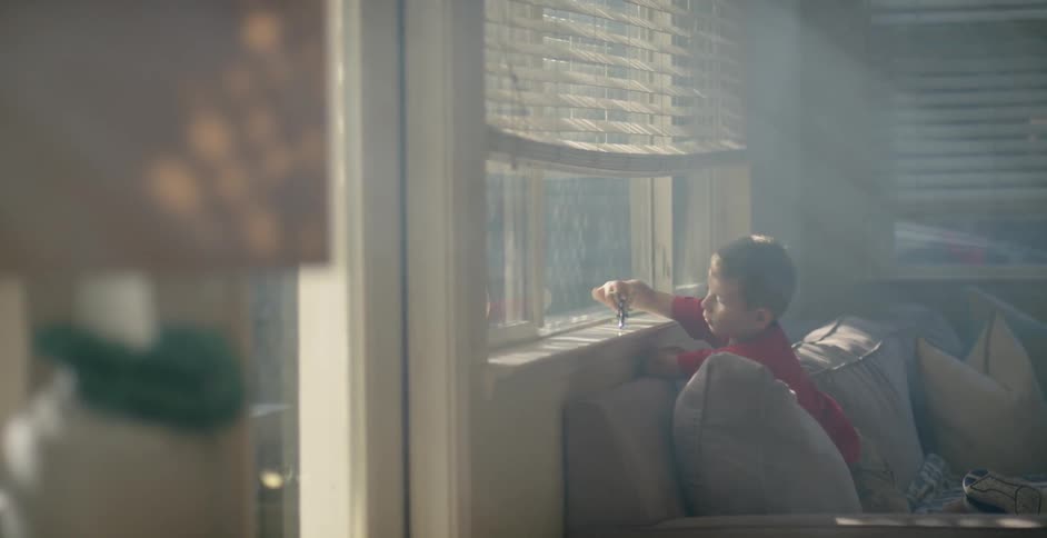 A boy leans on the back of a couch and plays with a toy on the window sill