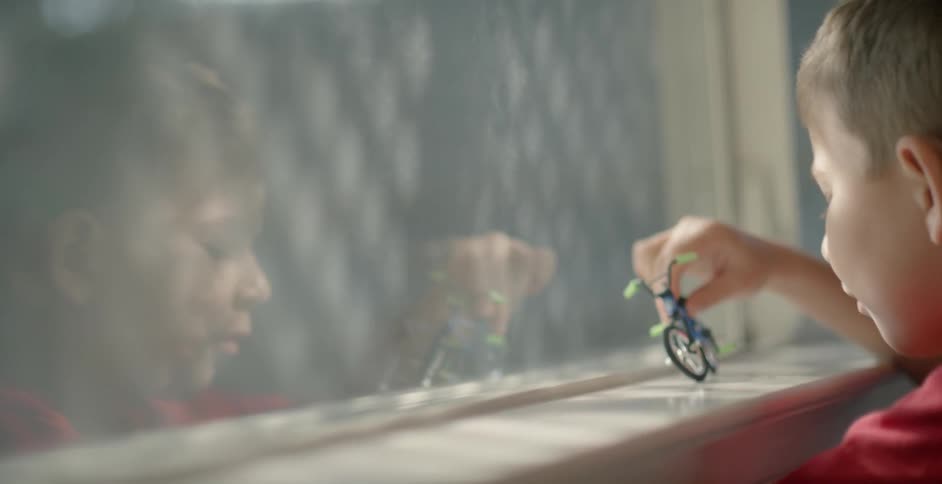 A boy playing with a toy bicycle on a window sill is reflected in the glass
