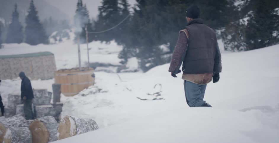 A man walks behind a rolling log in the snow towards two other men