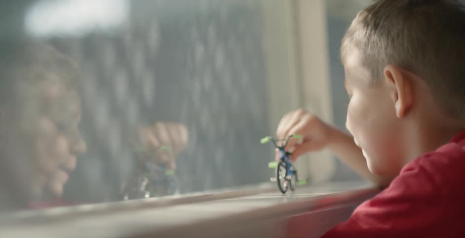 A boy plays with his toy bicycle on a window sill and is reflected in the glass