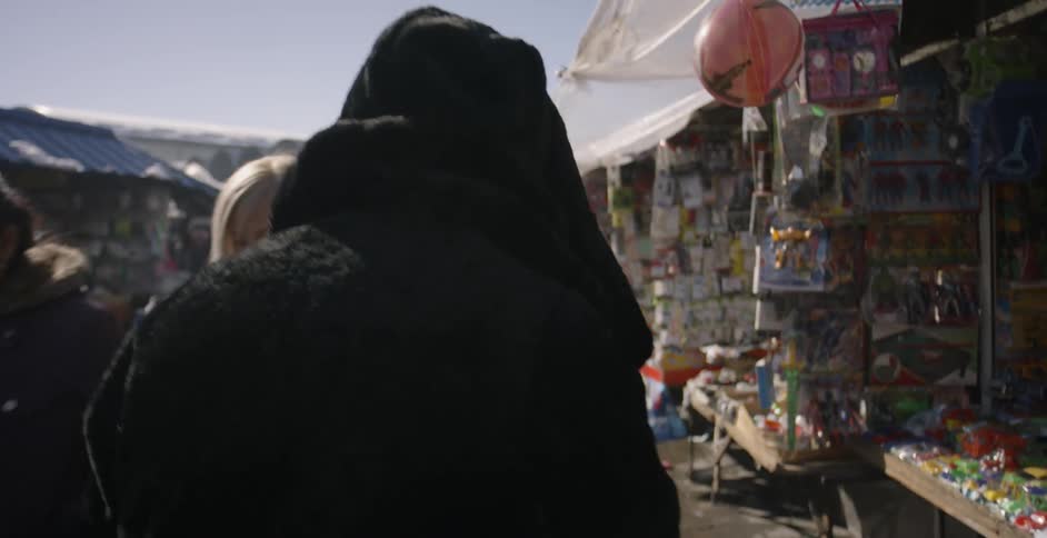 Person wearing a black coat and hood walks through a busy street market