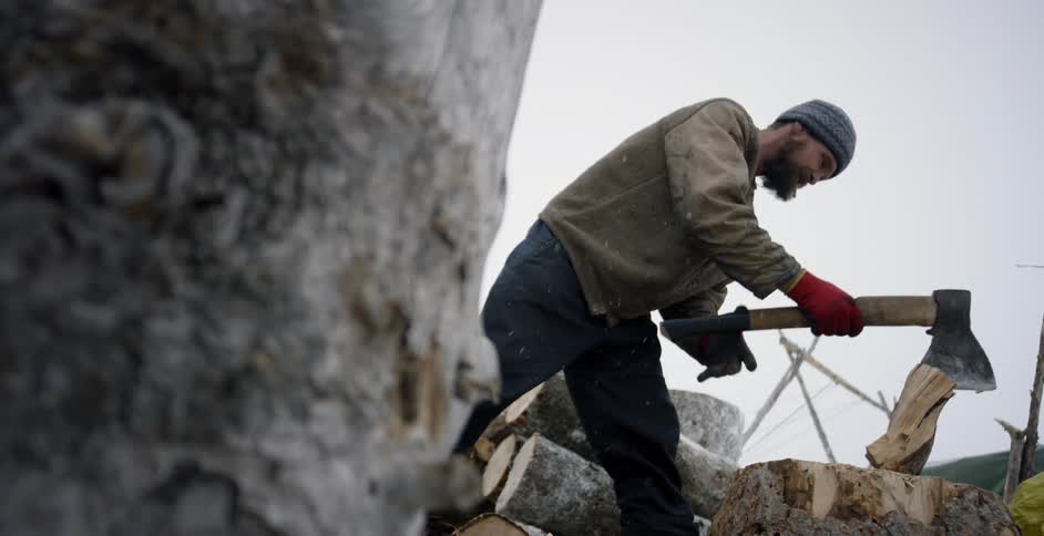 A man is using an ax to chop wood outside while it snows