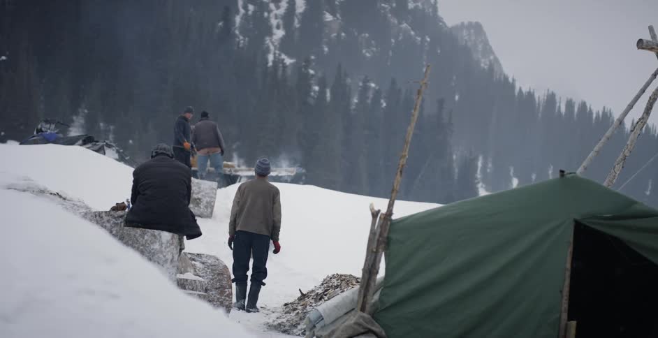Loggers rolling pieces of tree trunks down a snowy slope as others wait on them