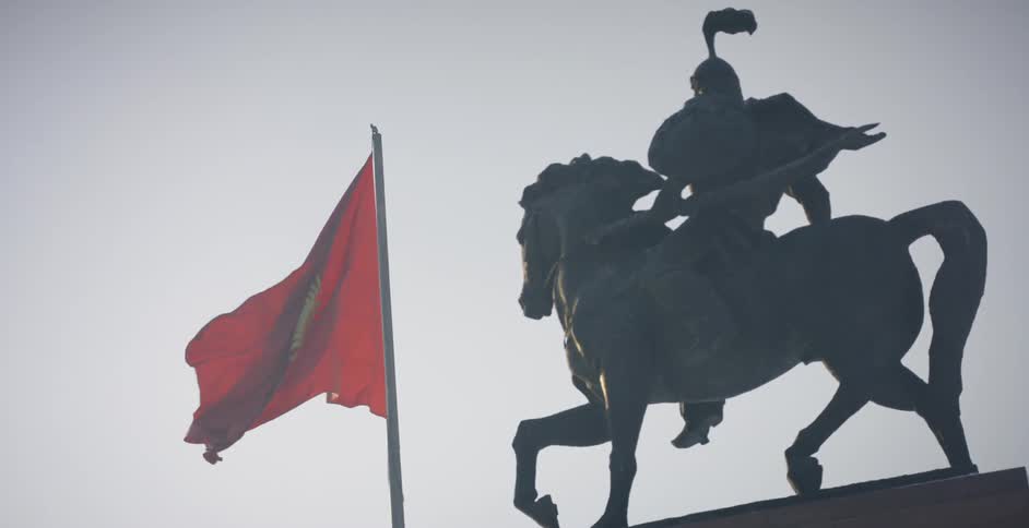 A kyrgyzstan flag is blowing in the breeze near a statue during the day