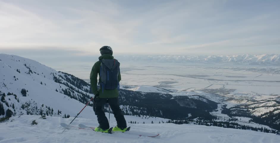 A skier is pausing to look at a view as he stands on a mountain slope in daylight