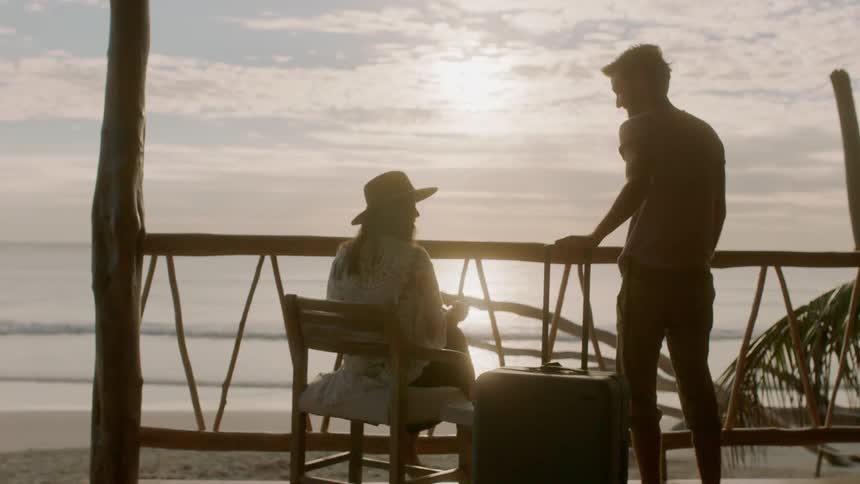 A woman who is reading a book is approached by a man pulling luggage