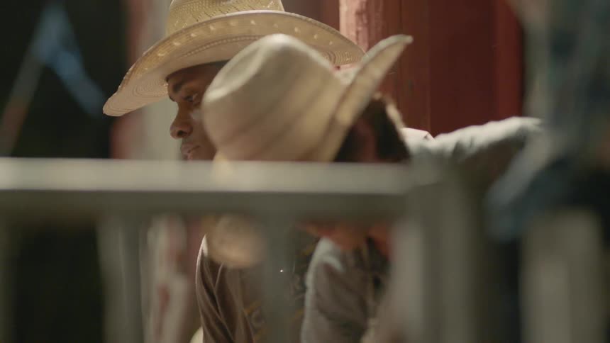 Two cowboys at a rodeo are hanging out on a fence during the day