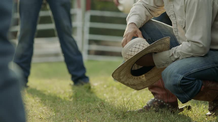A man leans towards the grass holding a cowboy hat