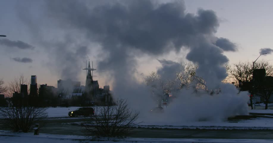 Clouds of steam are rising from the ground near a road in an urban area in winter