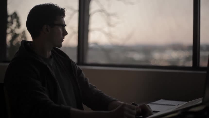 A man is leaning back in frustration as he writes at a desk at dusk