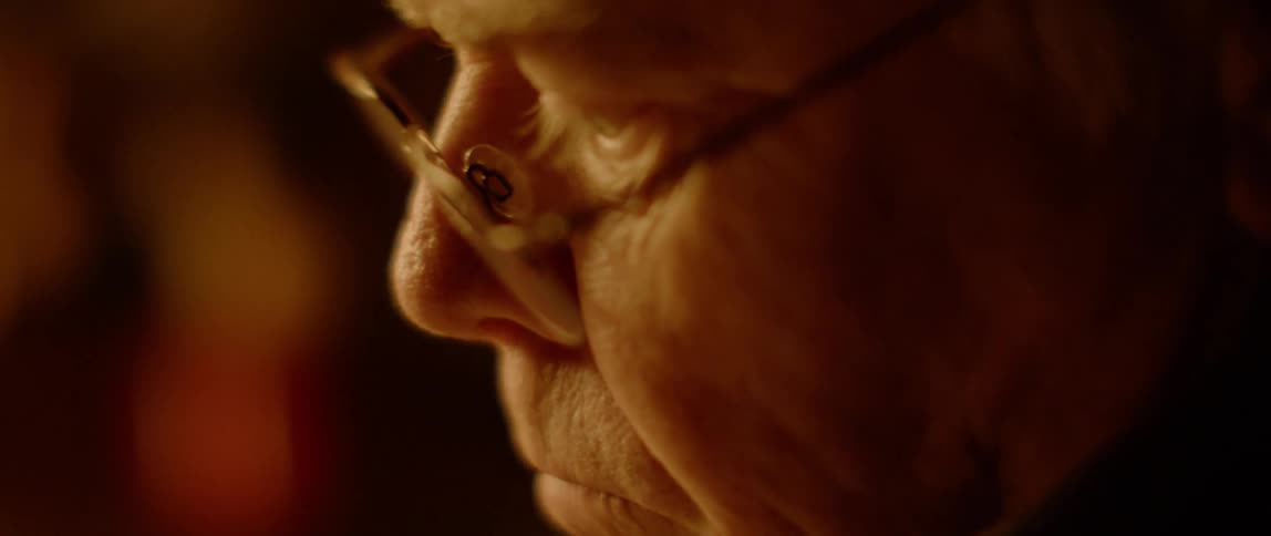 An elderly man wearing glasses looks downward