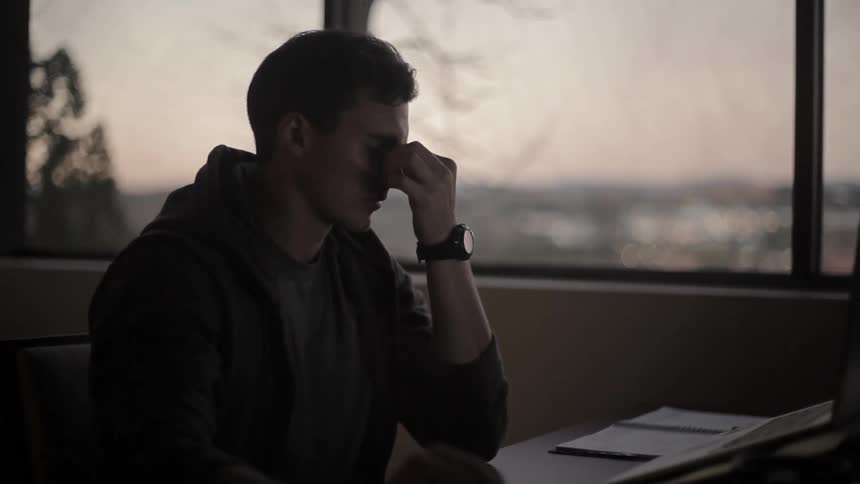 A man sitting at a desk working at dusk is removing his reading glasses