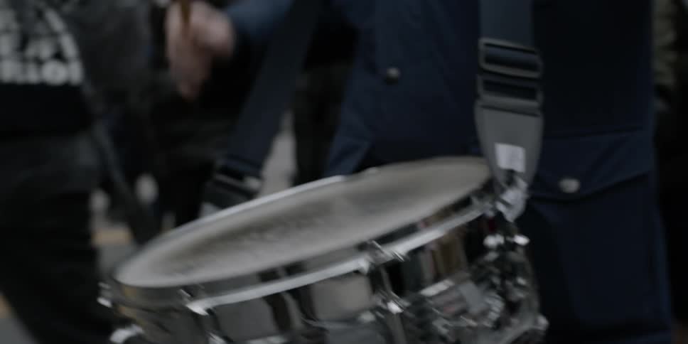 Man wears a drum on a strap around his neck and beats it with drumsticks