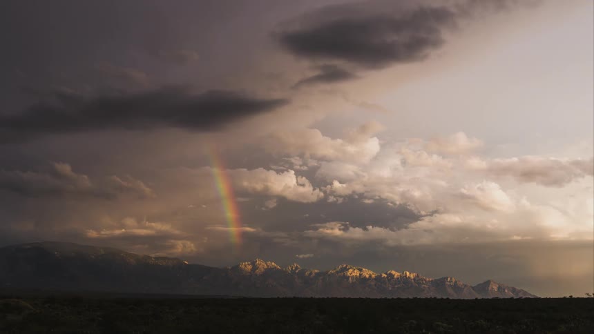 Clouds move over an expansive plain and mountains while unleashing a rainbow