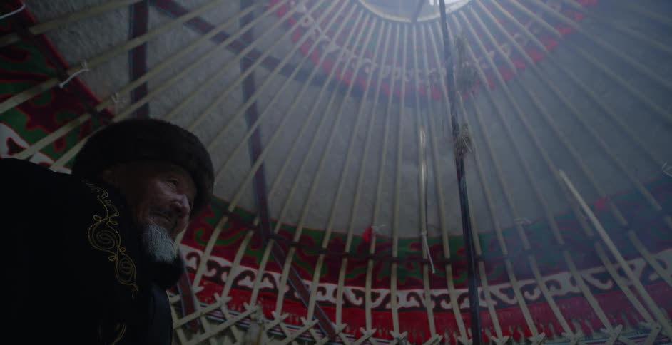 An elderly man sits in a yurt as smoke rises up to the opening