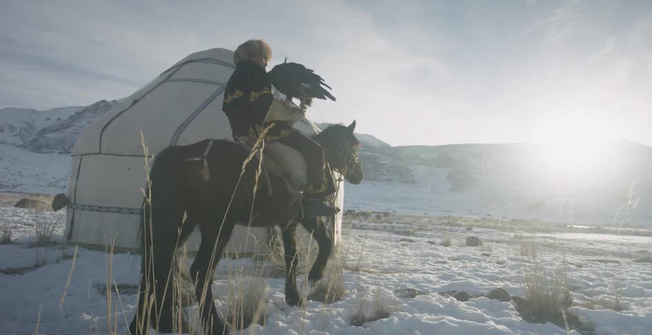 Mongolian men are riding horses past a yurt while carrying eagles in winter
