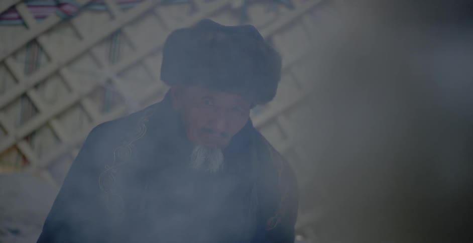 Smoke is billowing from a fire in front of an old man sitting in a yurt