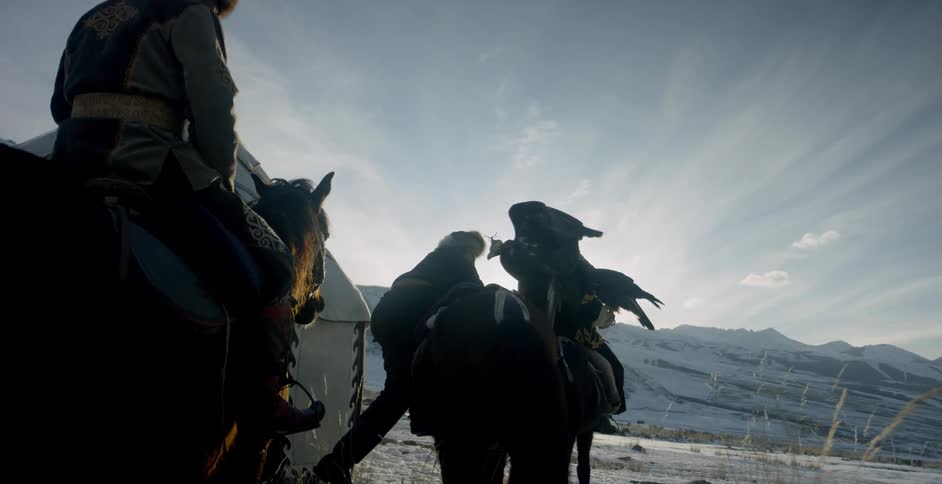 Mongolian men are riding horses past a yurt while carrying eagles in winter