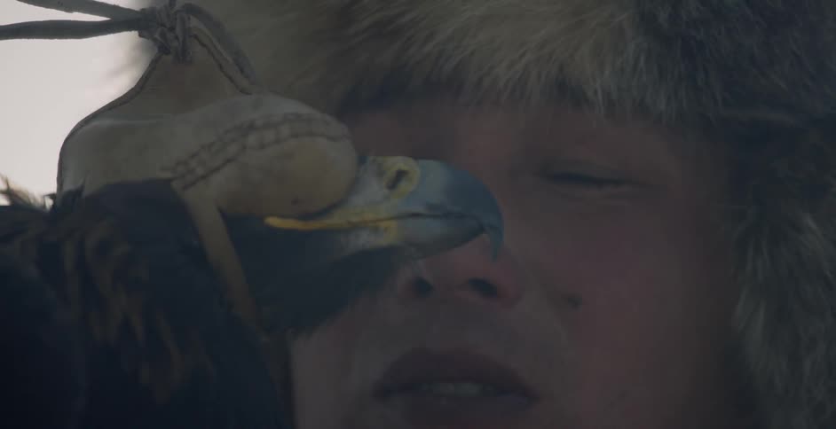 A mongolian man is holding an eagle outside in daylight