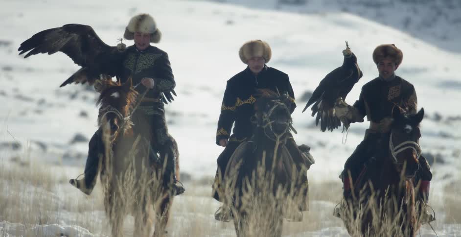 Men ride horses across snow with hunting eagles