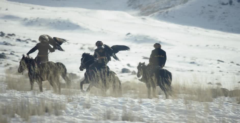 Three people ride horses in the snow and two carry birds on their arms