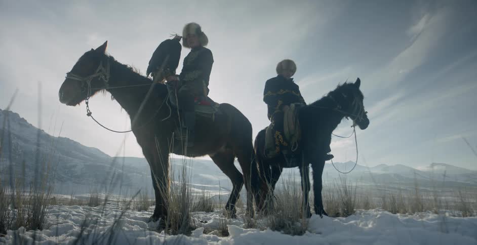 Two mongolian men are sitting on horses holding eagles outside in winter