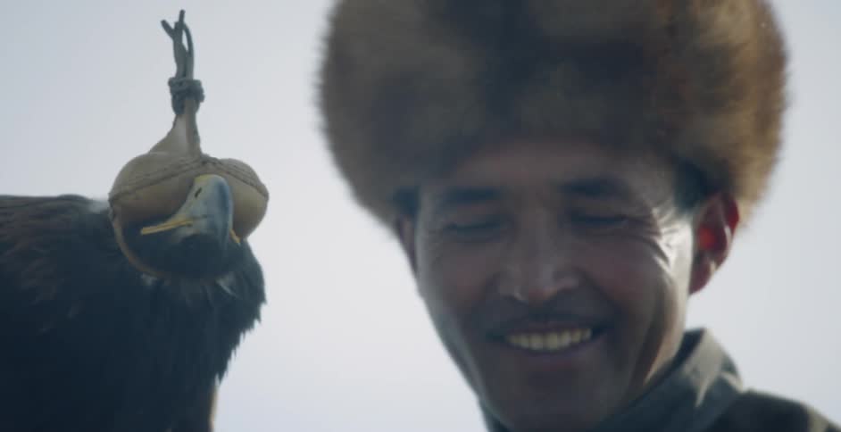 A mongolian hunter smiles while holding a hooded golden eagle