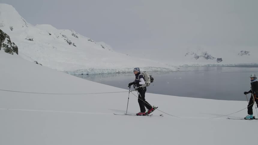 Two people walk on skis on the snow holding ski poles and a rope