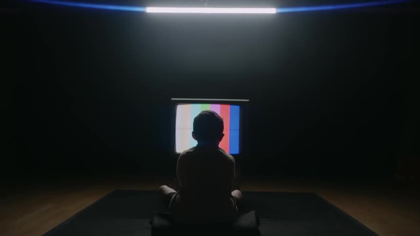 A boy sits on the floor in a dark room staring at a television