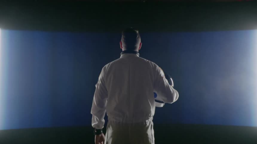 Man wearing a white suit holding a helmet looks at a large screen