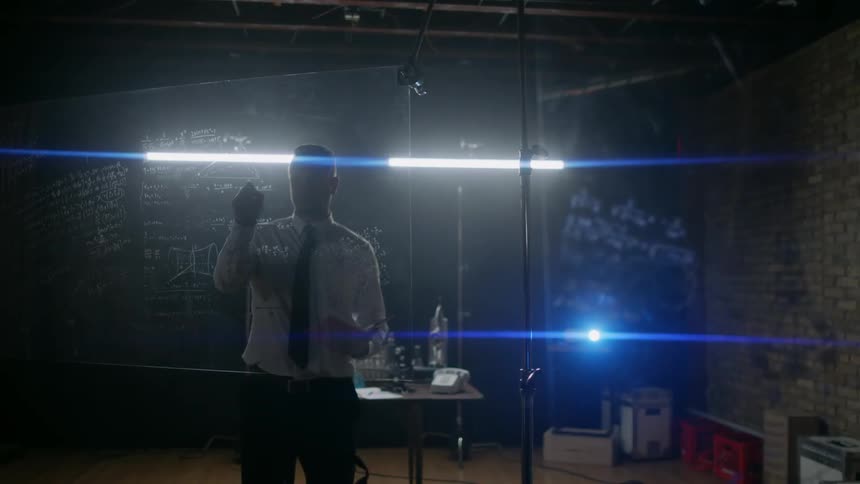 Man standing in a dark area writing on glass with some lights shining behind him