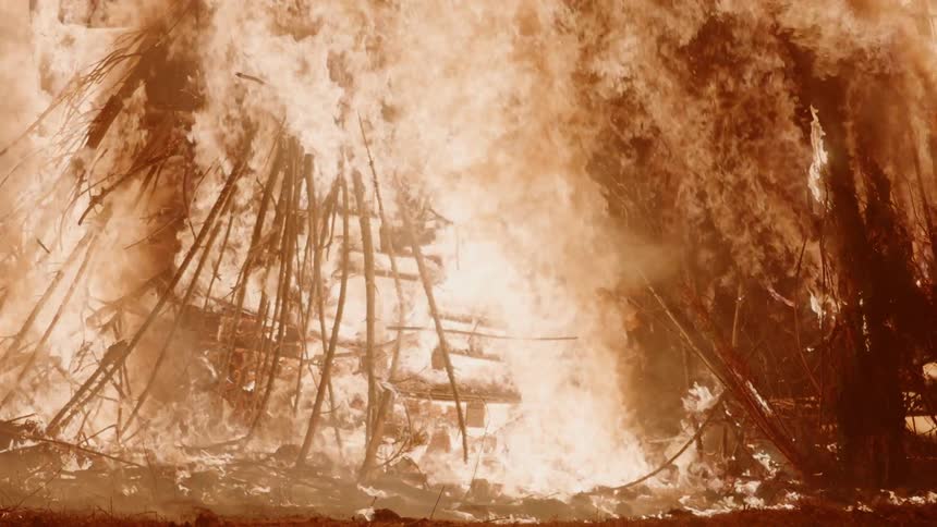 A bonfire is burning a structure of wood pallets and poles at night