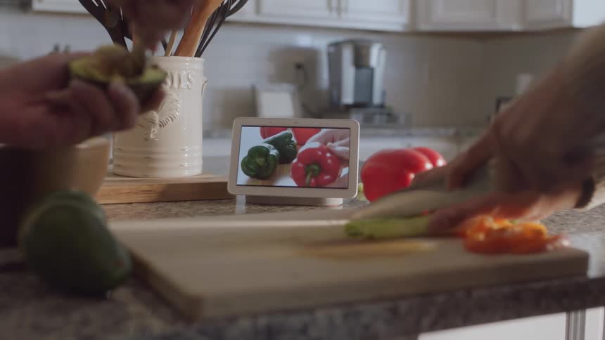 One person scoops out an avocado and another chops a vegetable on a cutting board by an instructional video