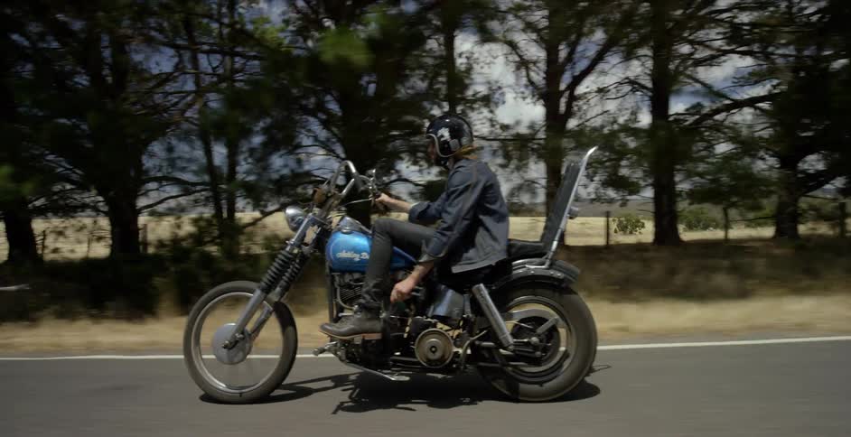Men ride motorcycles on a rural road past fields