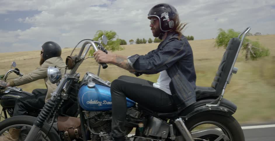 Two men ride motorcycles on a rural road on a sunny day
