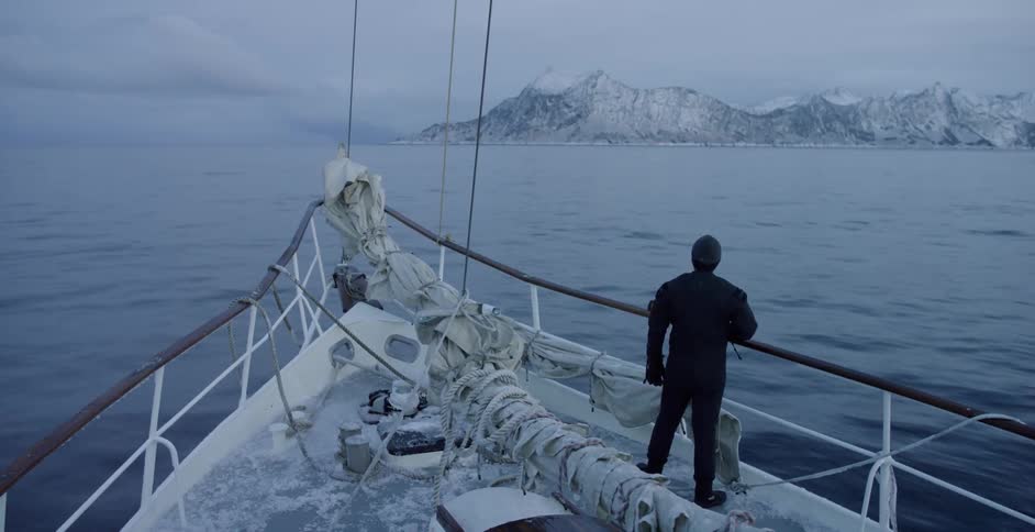 A man stands at the bow of a boat as it moves toward snow covered land