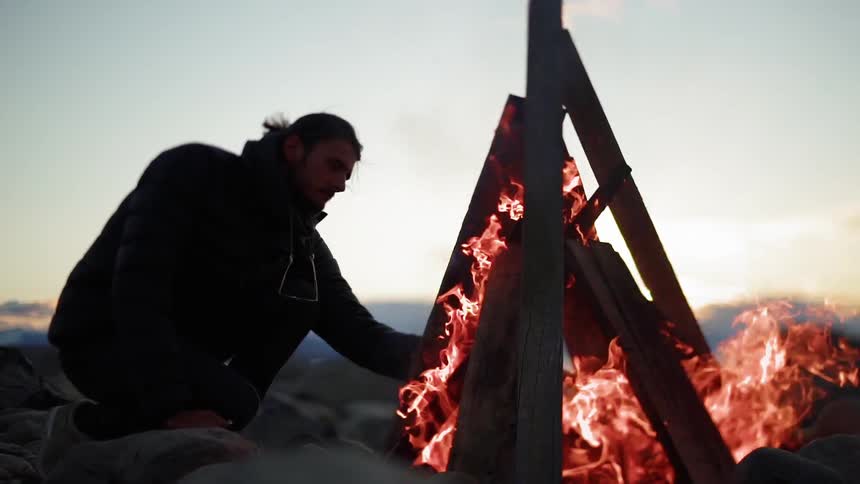 Man throws kindling on a blazing fire on the beach at sunrise