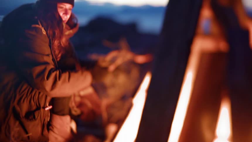 A woman is adding tinder to a bonfire outside at dusk