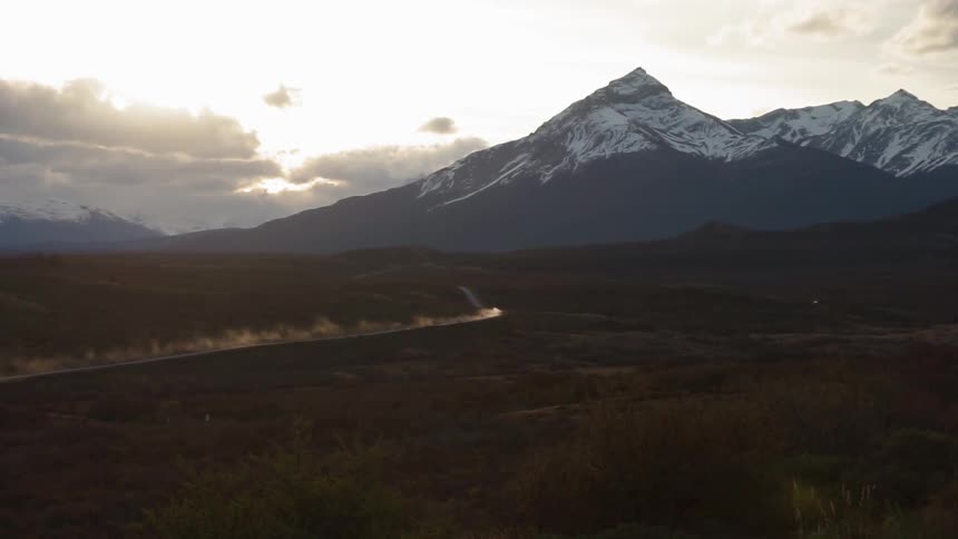 A vehicle kicks up a dirt trail as it drives on a road towards a mountain at dusk