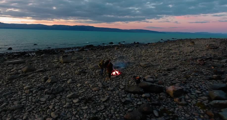 Two people stand next to a fire on a beach at sunset