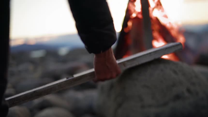 A man picks up a piece of wood and walks toward the fire