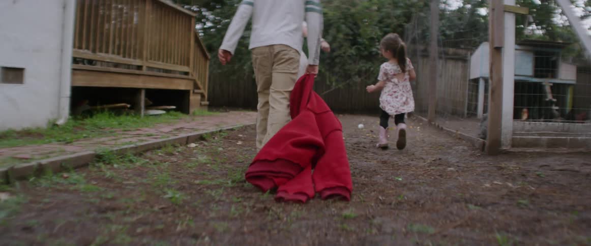 Two girls and a boy playing in a backyard as the boy drags his red jacket behind him
