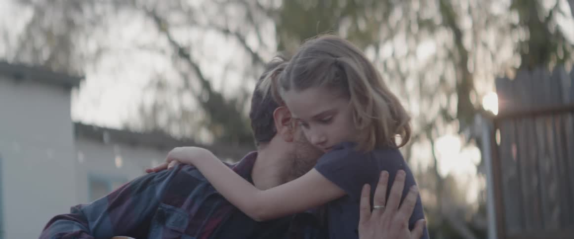A girl is hugging her father as he plays guitar in their backyard at dusk